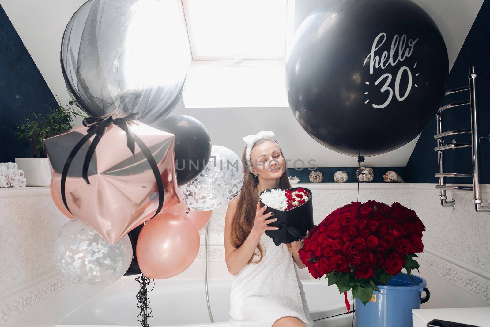 Portrait of positive beautiful brunette woman in headband and towel enjoying flowers in box surrounded by colorful air balloons and bouquet of red roses. She is in the bathroom recieving presents.