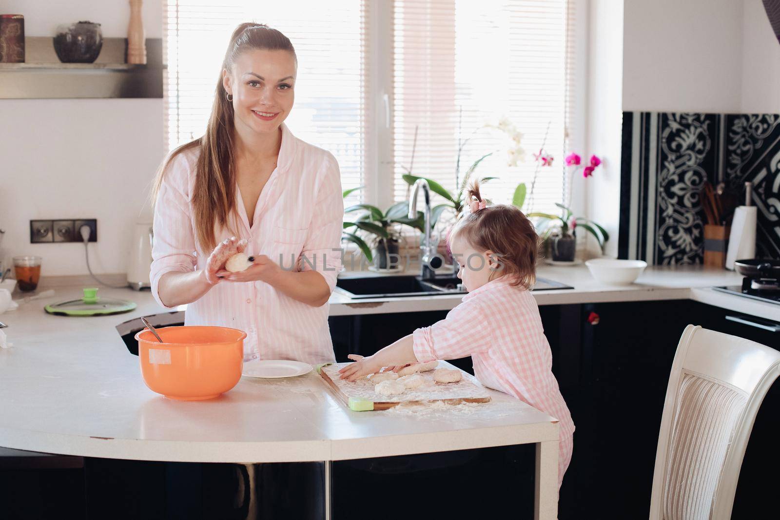 Happy wife smiling and preparing breakfast together with child for husband. Beautiful mother with cute daughter cooking in pajamas in morning. Little lovely kid helping parent with dough at kitchen.