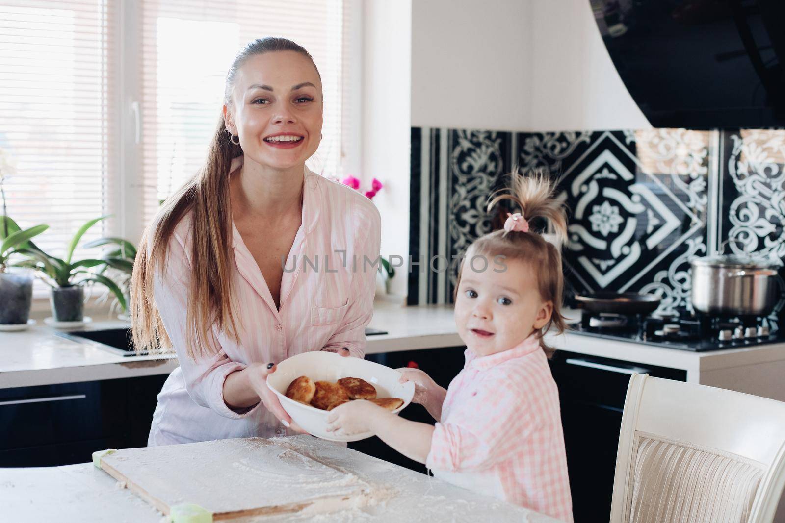 Beautiful woman with lovely child in pajamas holding big bowl with cookies. Close up of happy mother and daughter sitting together at kitchen in morning. Little kid helping parent cooking breakfast.