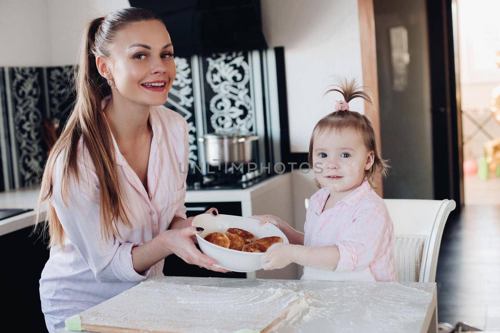 Beautiful woman with lovely child in pajamas holding big bowl with cookies. Close up of happy mother and daughter sitting together at kitchen in morning. Little kid helping parent cooking breakfast.