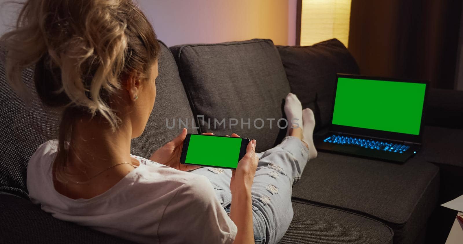 Over the shoulder view woman lying on a sofa using phone and laptop with Green Mock-up Screen Chroma key watching content. Horizontal phone Green Screen. Using smartphone home.