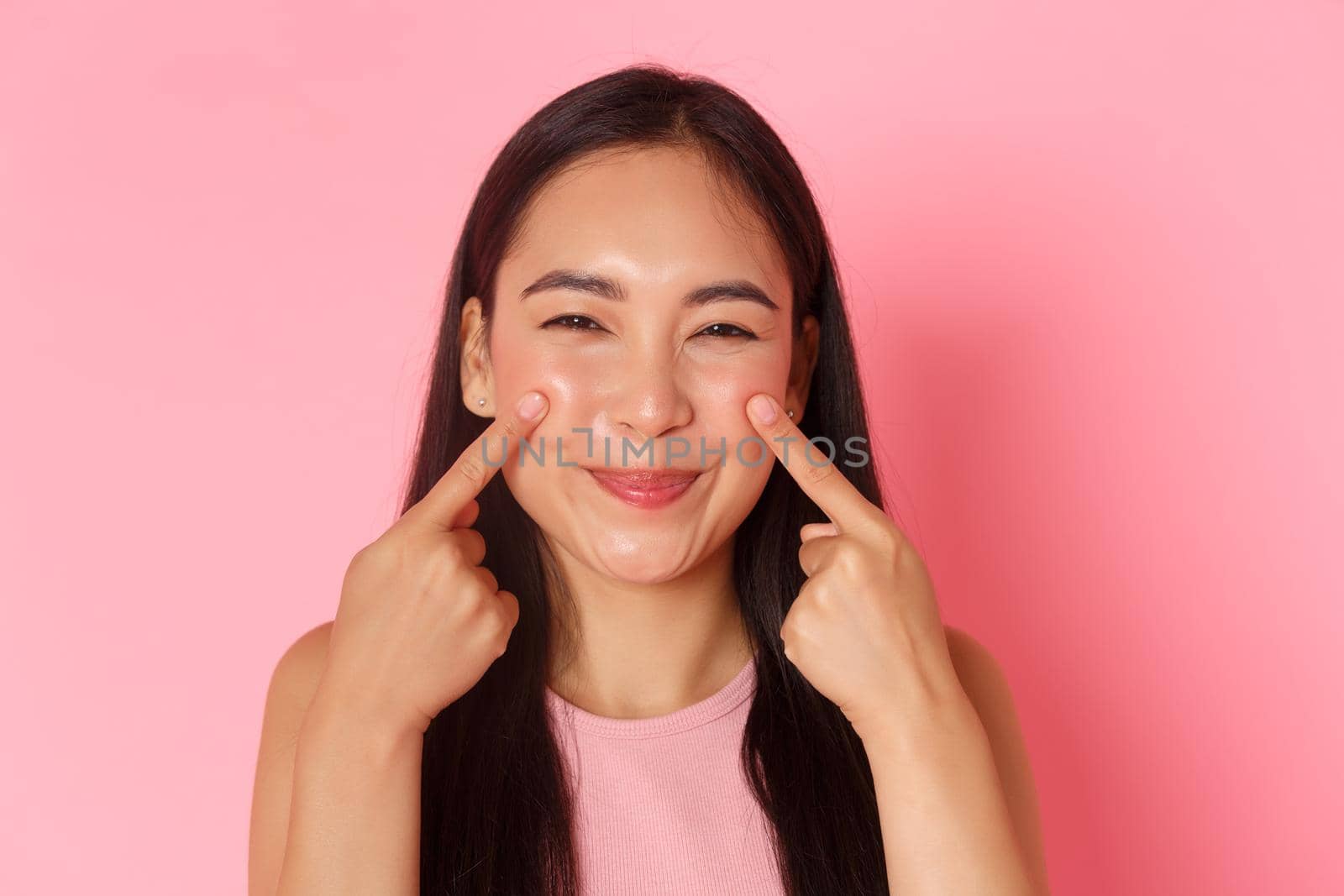 Beauty, fashion and lifestyle concept. Close-up of silly and cute, adorable asian girl poking her cheeks and smiling happily, promo of skincare or makeup product, pink background.