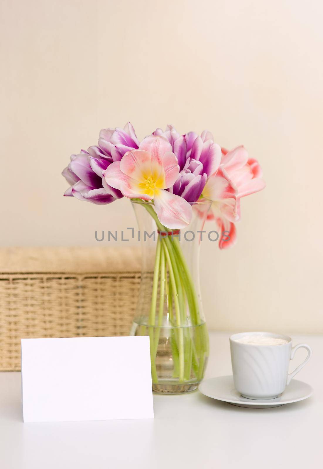 A bouquet of opened pink and purple tulips, a wicker basket, a white cup and an empty postcard with space for text, on a white table in a bright room. Vertical