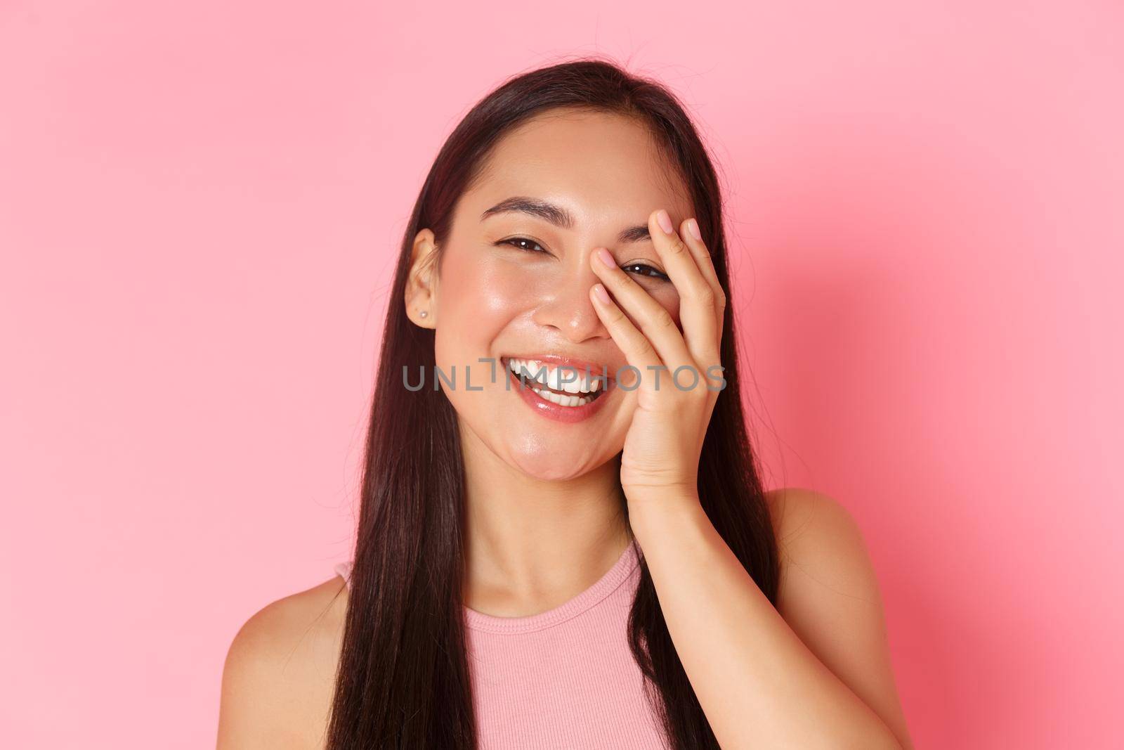 Beauty, fashion and lifestyle concept. Close-up of carefree beautiful asian girl touching face and laughing happily with closed eyes, standing over pink background joyful, skincare product promo by Benzoix