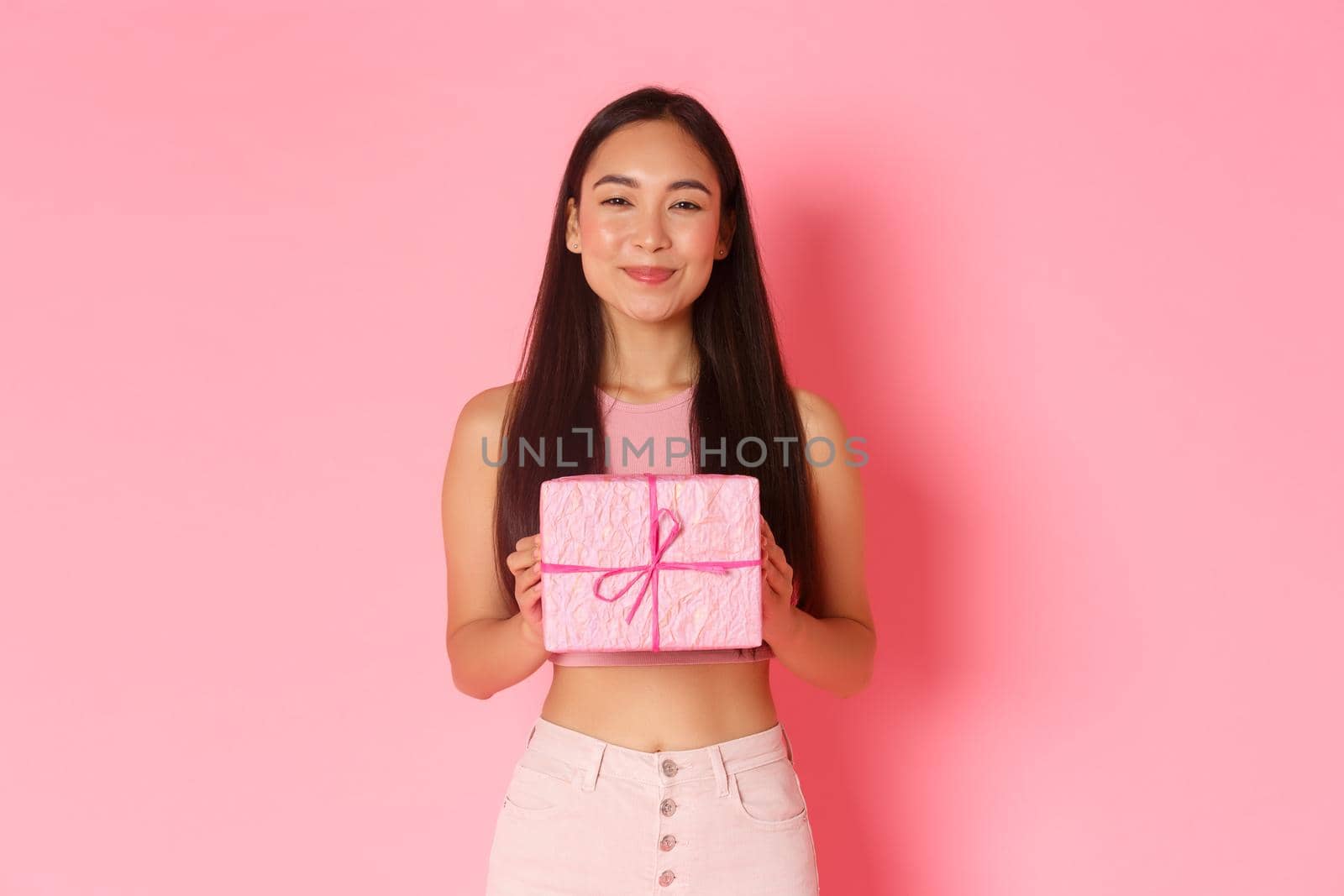 Holidays, celebration and lifestyle concept. Close-up of pleased cheerful asian girl receive cute wrapped gift and smiling satisfied, likes receiving presents, standing over pink background by Benzoix