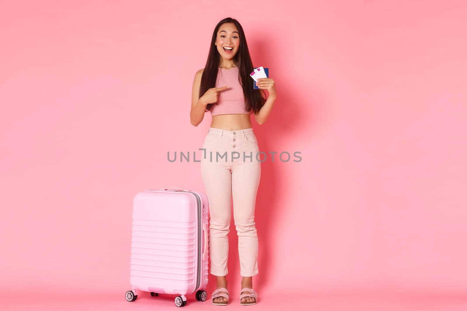 Travelling, holidays and vacation concept. Full-length of happy smiling asian girl tourist standing near suitcase, pointing finger at airline flight tickets and passport, pink background.