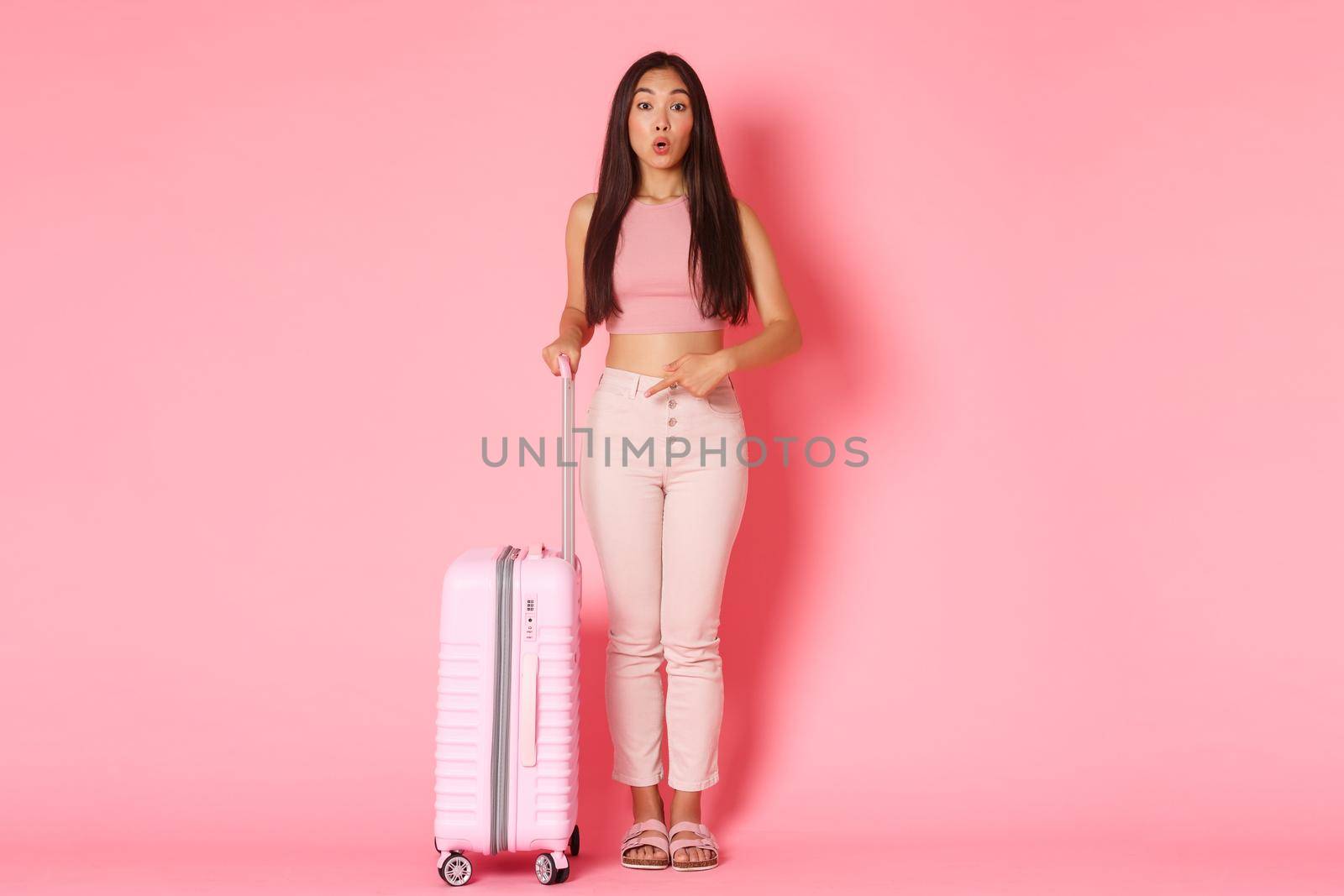 Travelling, holidays and vacation concept. Full-length of pretty surprised girl tourist folding lips amazed as pointing finger at suitcase, impressed with how much staff it can hold, pink background by Benzoix