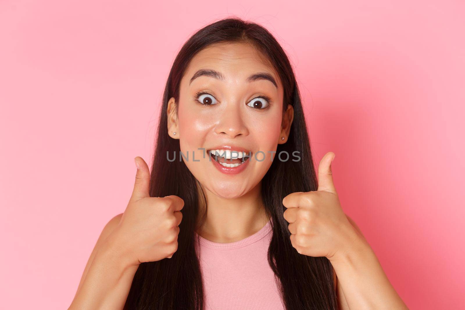 Beauty, fashion and lifestyle concept. Close-up of enthusiastic good-looking asian girl like idea, praising great choice, looking amazed and surprised showing thumbs-up in approval, pink wall by Benzoix