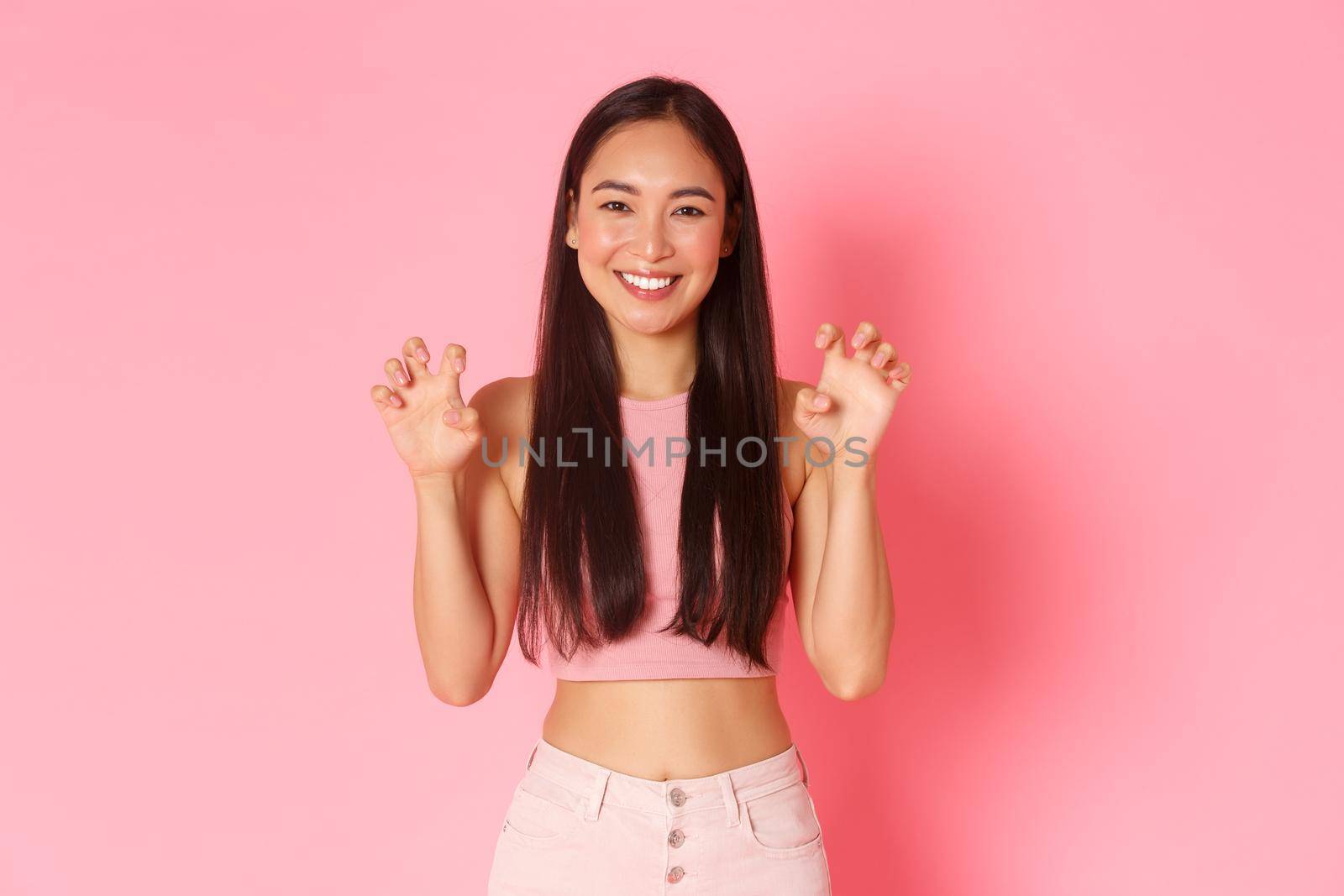 Portrait of silly and cute, upbeat asian girl in trendy summer clothes having fun, acting like lion, making roar gesture with hands mimicking paws, standing over pink background smiling.