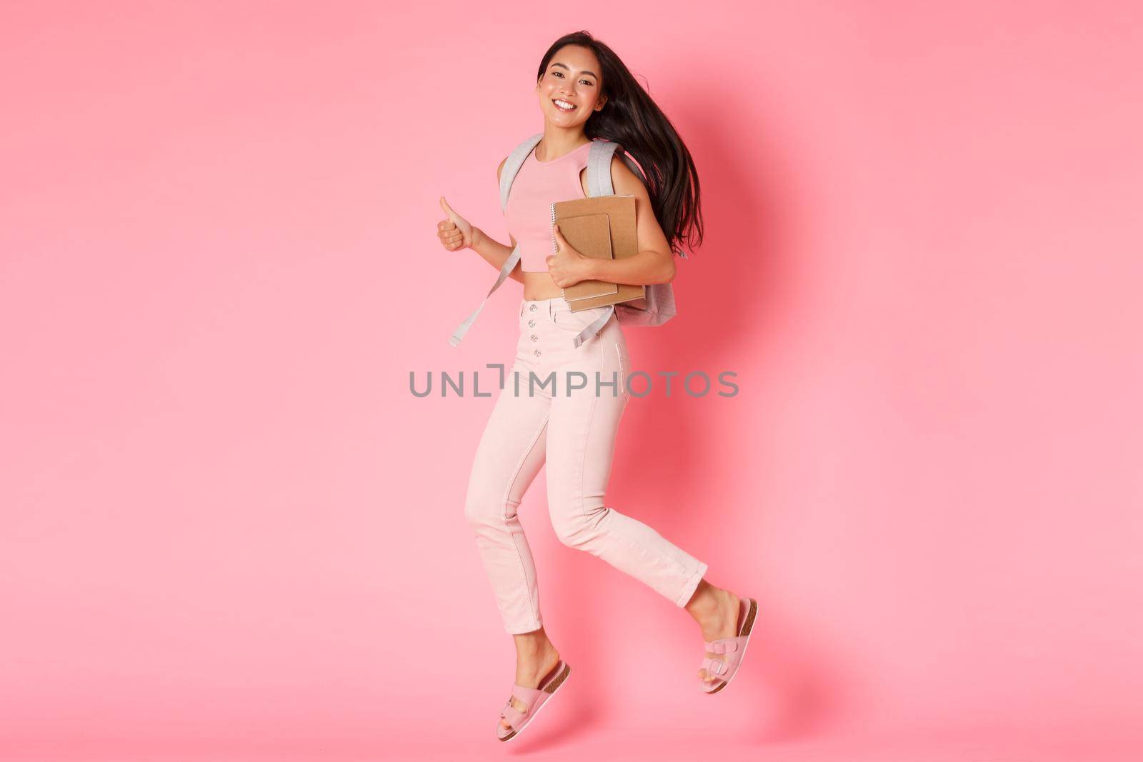 Back to school, education and lifestyle concept. Full length of happy smiling asian female student, girl from college jumping with notebooks and backpack, showing thumbs-up in approval, pink wall.