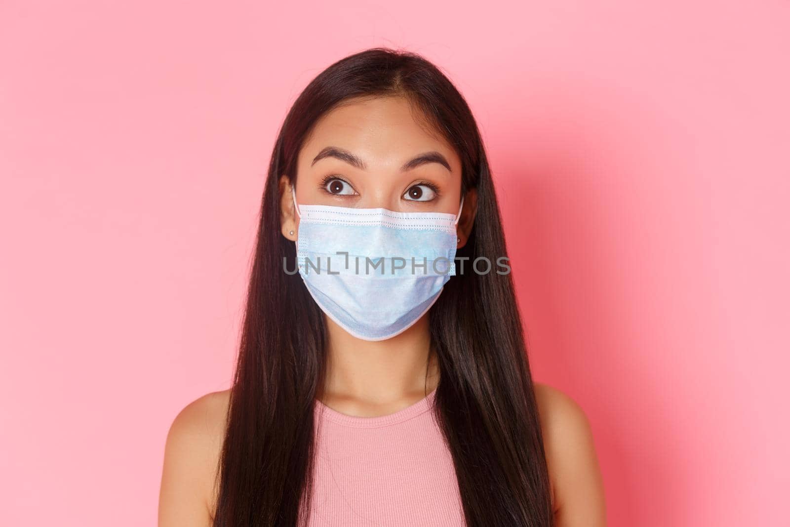 Covid-19 pandemic, coronavirus and social distancing concept. Close-up of thoughtful and curious asian girl in medical mask found something interesting, looking upper left corner, pink background.