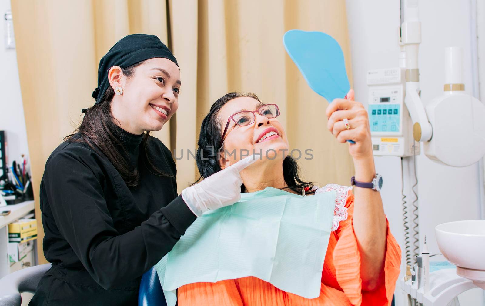 Satisfied female client in dental clinic looking at mirror, Dentist with patient smiling at hand mirror in office, female patient checking teeth after curing teeth in dental clinic, by isaiphoto
