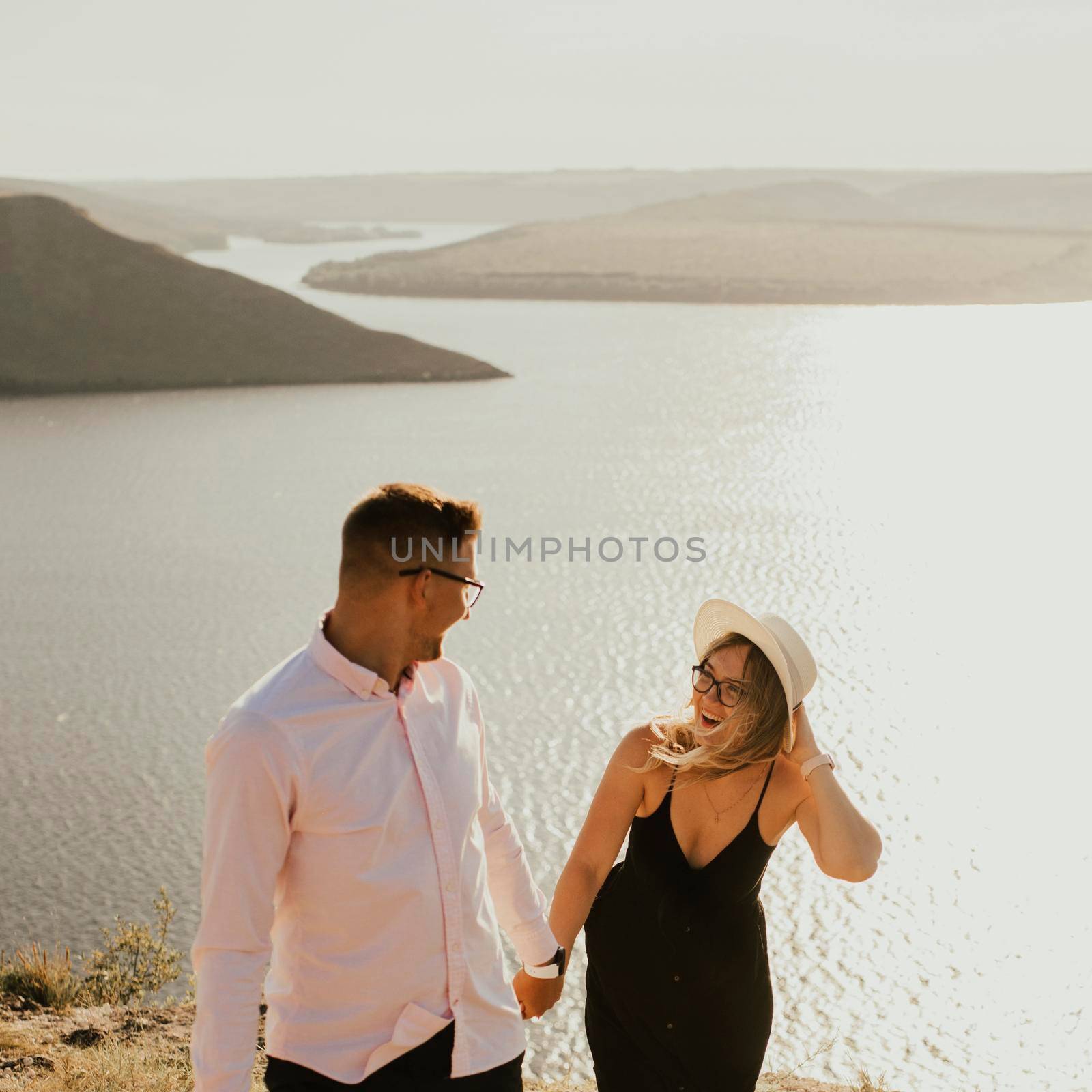 A man and a woman in love walk on a mountain above a large lake by the sea at sunset. A couple of fair-haired fair-skinned people in love are resting in nature in a field