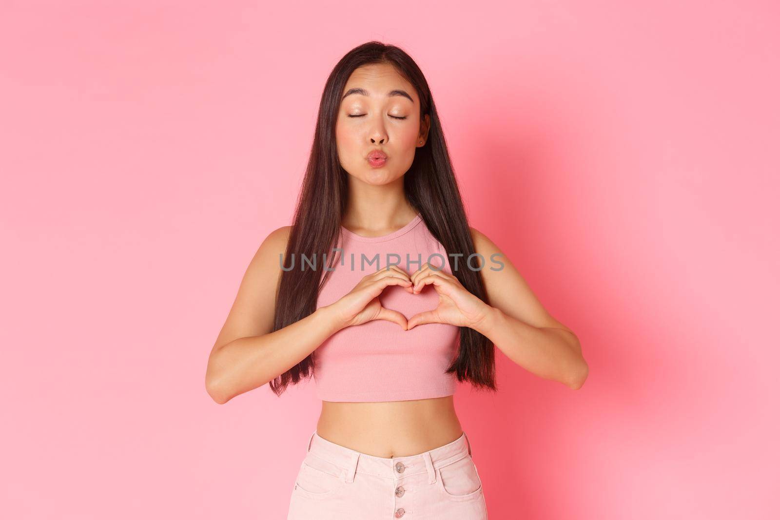 Lifestyle, beauty and women concept. Portrait of romantic beautiful asian girl daydreaming, fell in love, making heart gesture over chest, close eyes and pouting for kiss, standing pink background.