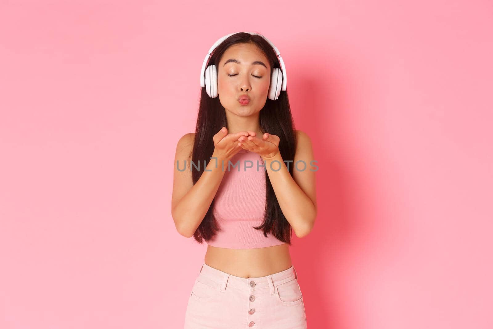 Waist-up portrait of silly and flirty asian girl sending air kiss at camera with closed eyes while listening music in headphones, passionate girl make mwah to camera, pink background.