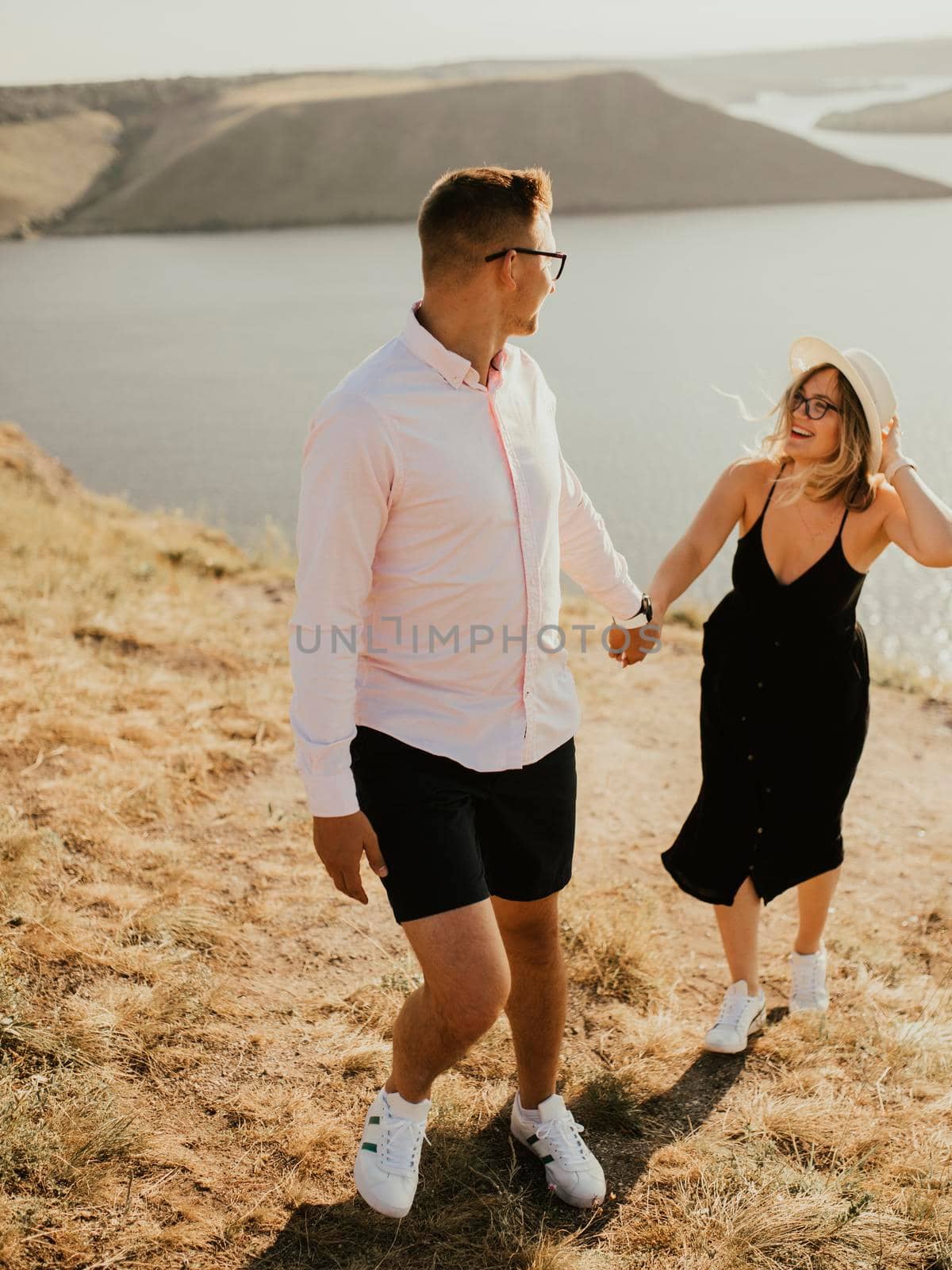 A man and a woman in love walk on a mountain above a large lake by the sea at sunset. A couple of fair-haired fair-skinned people in love are resting in nature in a field