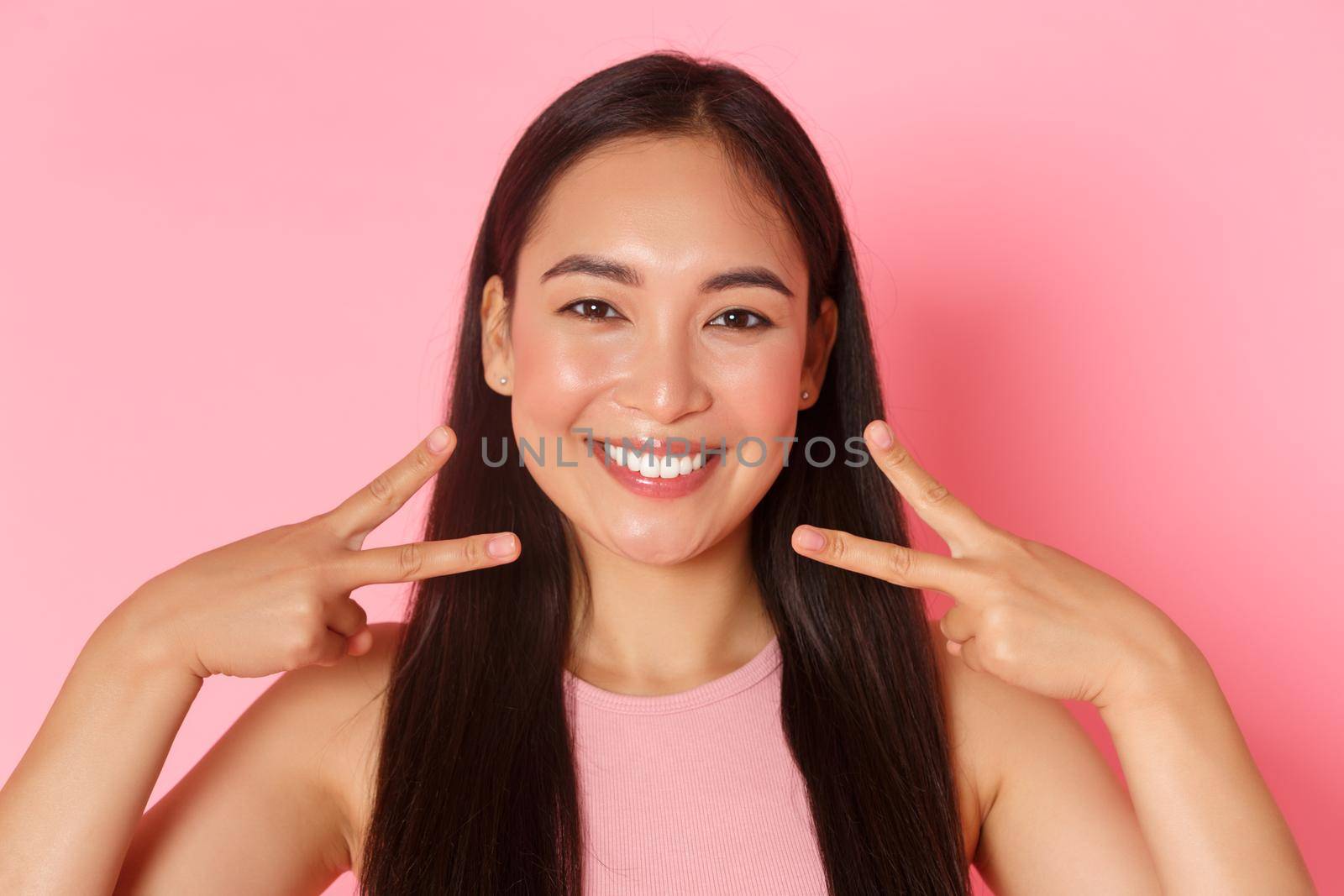 Beauty, fashion and lifestyle concept. Close-up of beautiful glamour asian girl with white perfect smile, clean face, showing peace kawaii gesture and looking happy, pink background.