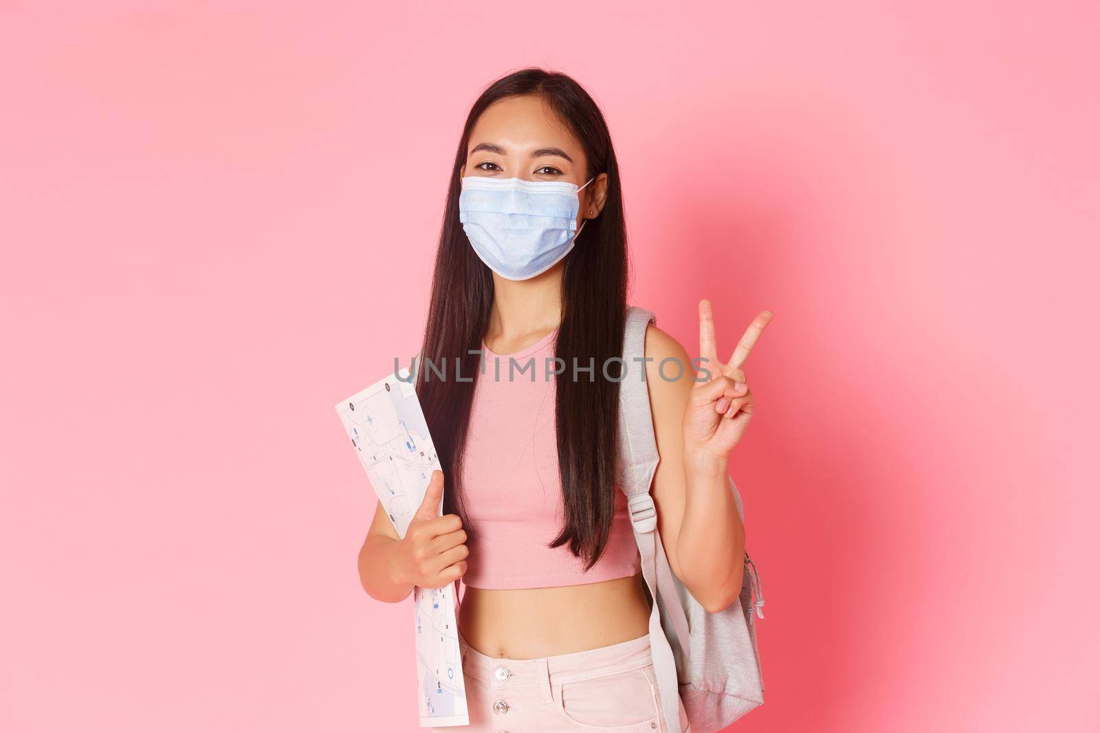 Safe tourism, travelling during coronavirus pandemic and preventing virus concept. Happy asian girl travel in pandemic covid-19 with medical mask, holding map, show peace sign, tourist going abroad by Benzoix