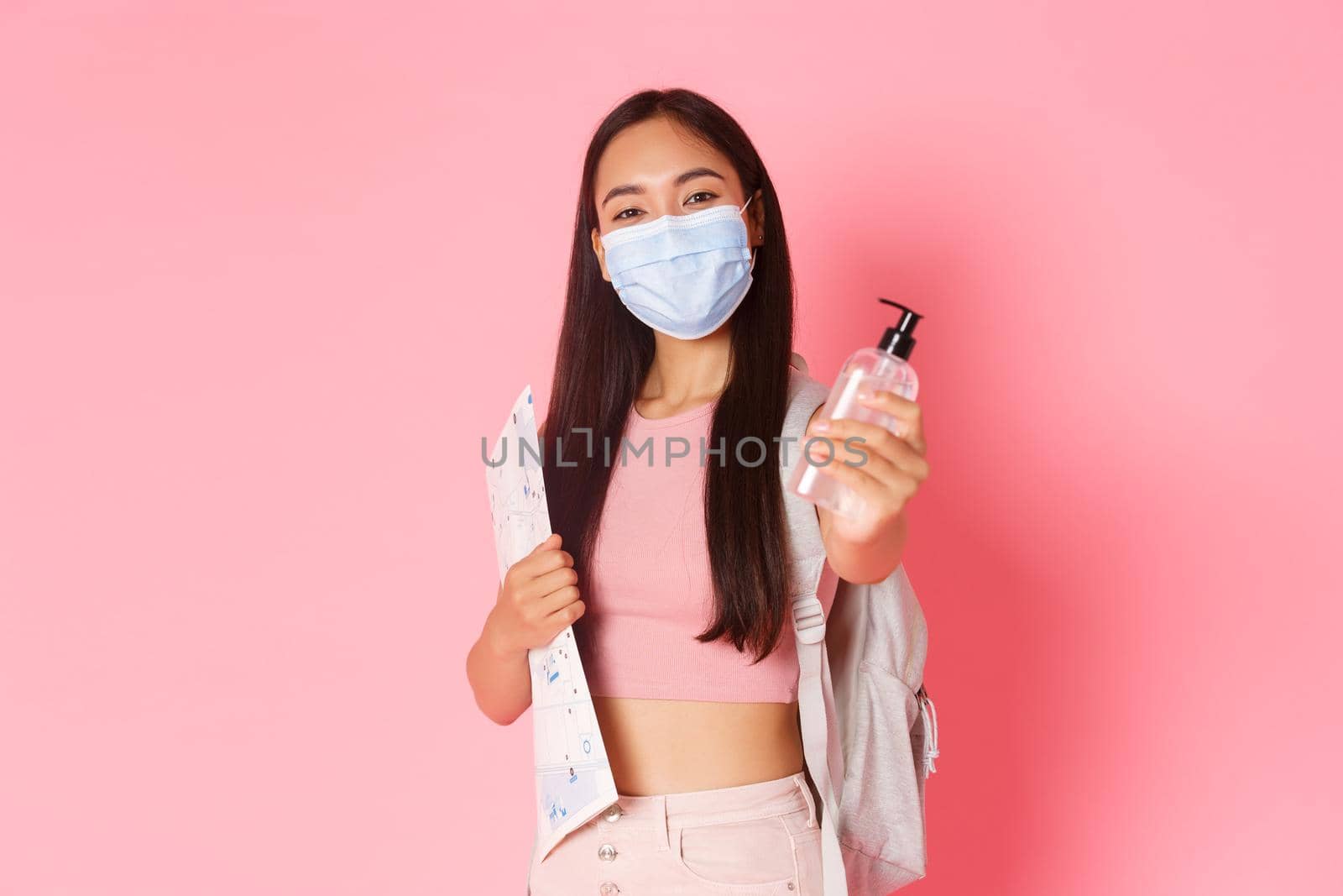 Safe tourism, travelling during coronavirus pandemic and preventing virus concept. Cute asian girl tourist on vacation with medical mask and hand sanitizer, holding map of city, pink background by Benzoix