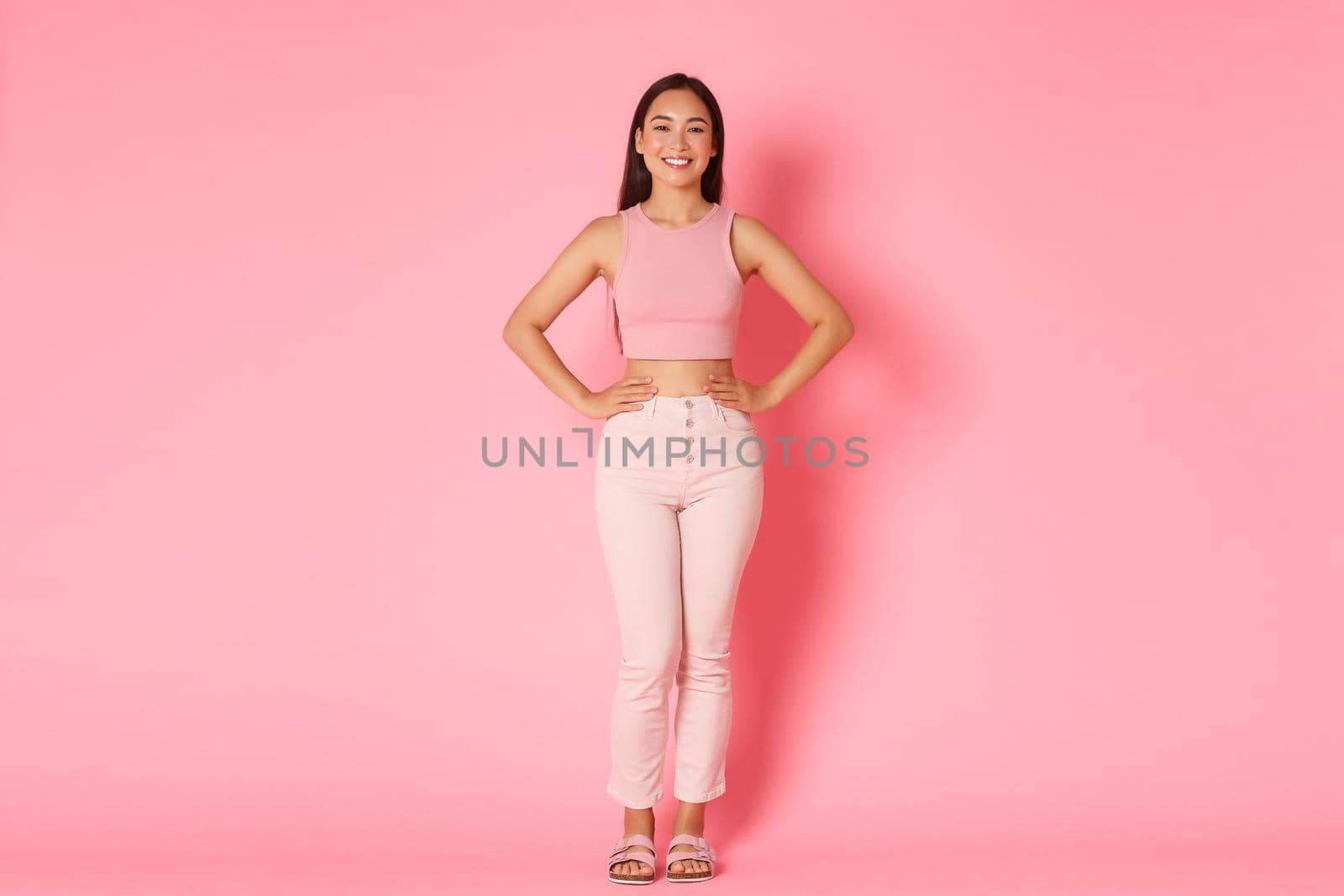 Full length portrait of fashionable brunette asian girl standing over pink background with sassy smile, holding hands over waist confident. Korean woman picking new outfit, shopping in malls.