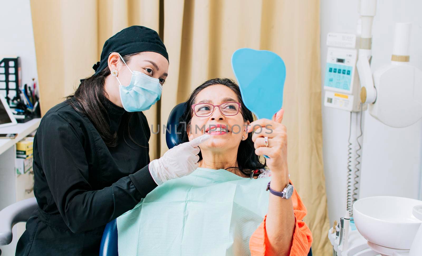 Satisfied female client in dental clinic looking at mirror, Dentist with patient smiling at hand mirror in office, female patient checking teeth after curing teeth in dental clinic, by isaiphoto