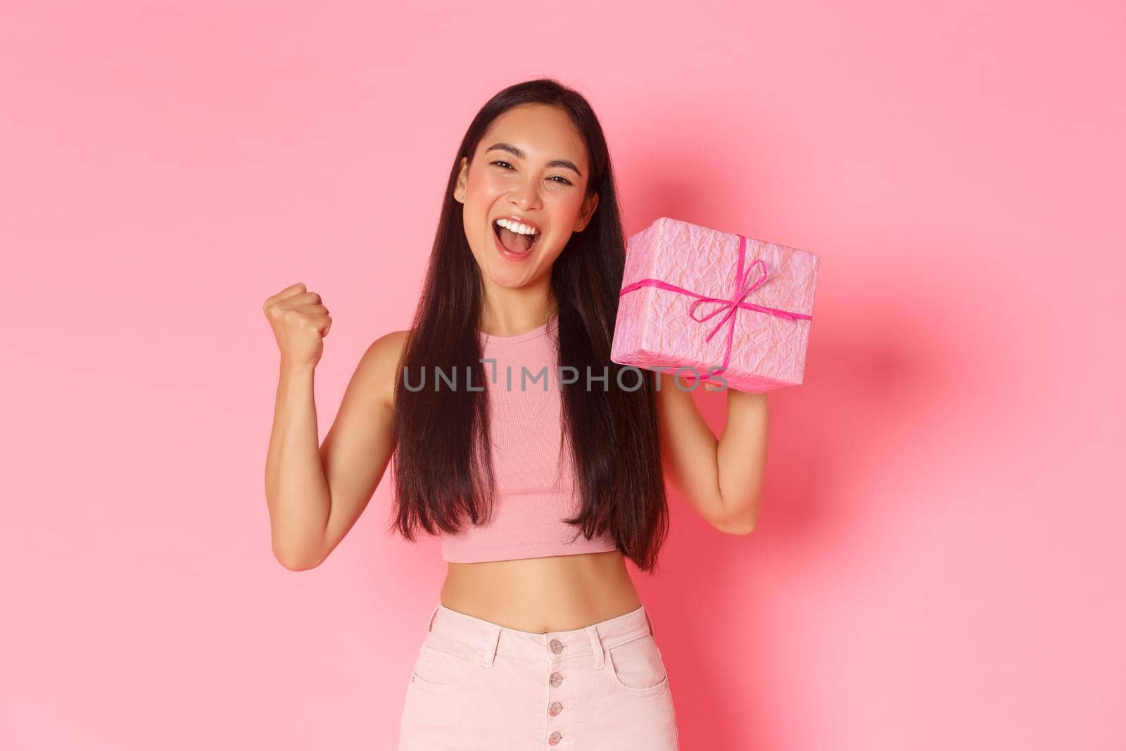 Holidays, celebration and lifestyle concept. Triumphing happy asian cute birthday girl looking upbeat, likes receiving gifts, raising fist pump and showing wrapped present, standing pink background by Benzoix