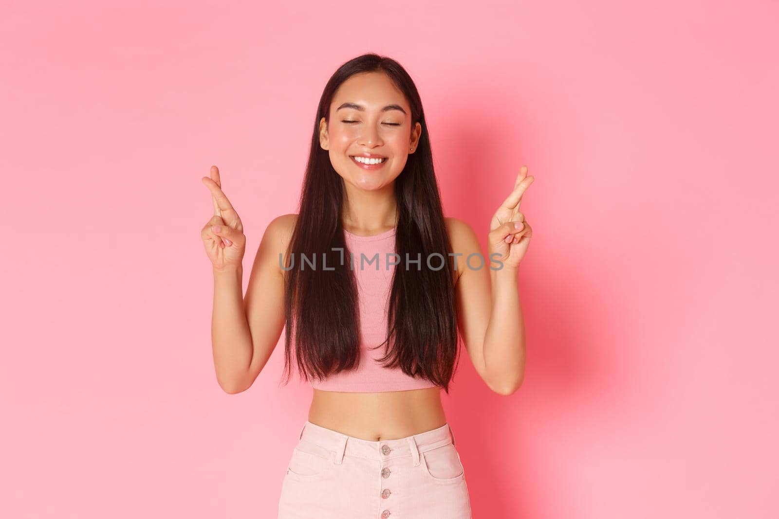 Lifestyle, beauty and women concept. Portrait of hopeful, happy asian girl in summer clothes making wish, cross fingers good luck smile and close eyes, praying over pink background by Benzoix