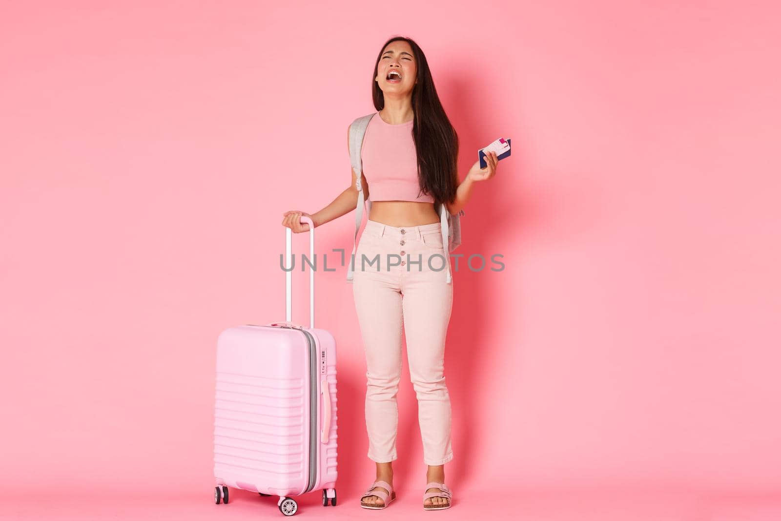 Travelling, holidays and vacation concept. Full-length of crying, sad and distressed asian girl tourist, feeling depressed about missed flight, standing suitcase and plane tickets, pink background.