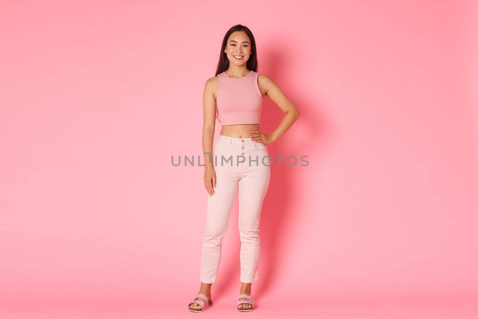 Full-length of stylish feminine korean girl in glamour outfit, holding hand on waist and smiling sassy at camera, standing over pink background. Concept of shopping, beauty and fashion.