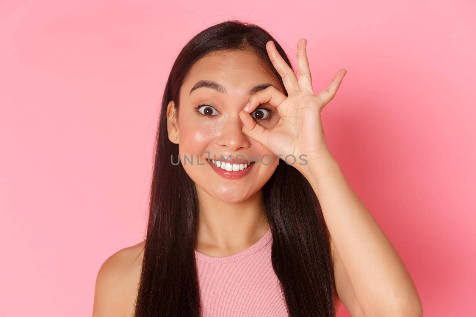 Beauty, fashion and lifestyle concept. Close-up of dreamy and cute asian young girl looking through okay gesture or showing zero, smiling upbeat, found something perfect, pink background.