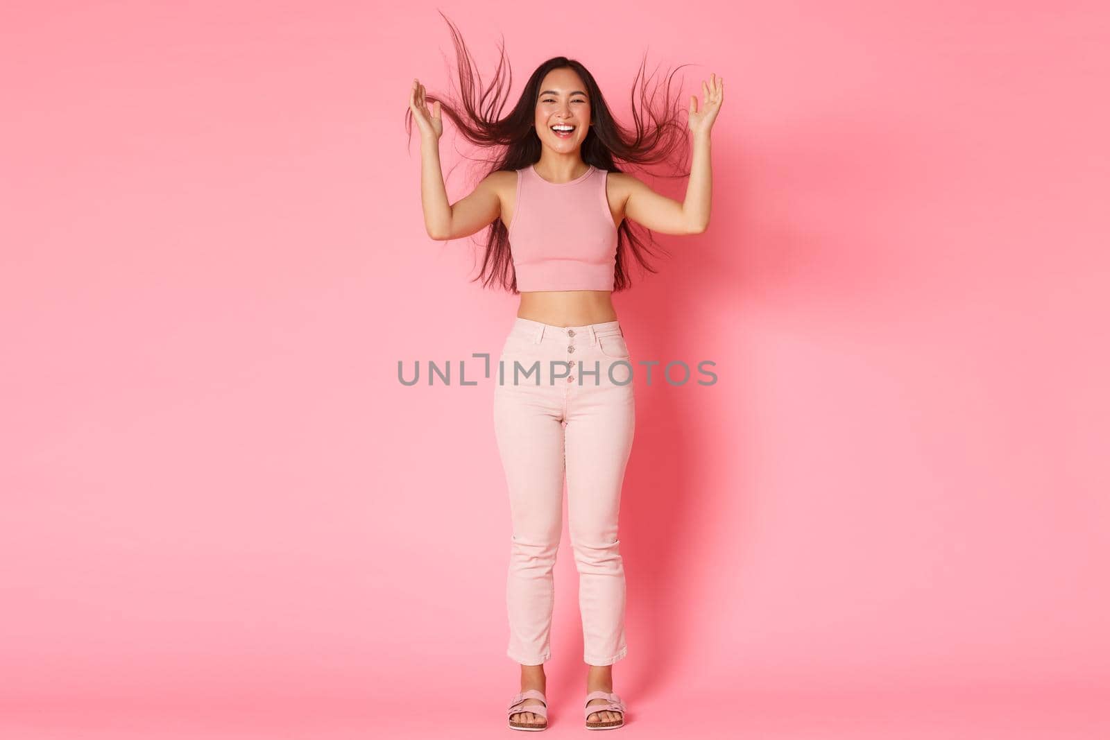 Fashion, beauty and lifestyle concept. Full length of gorgeous upbeat brunette asian girl tossing hair and laughing carefree, wearing glamour clothes, promote haircare products over pink background.