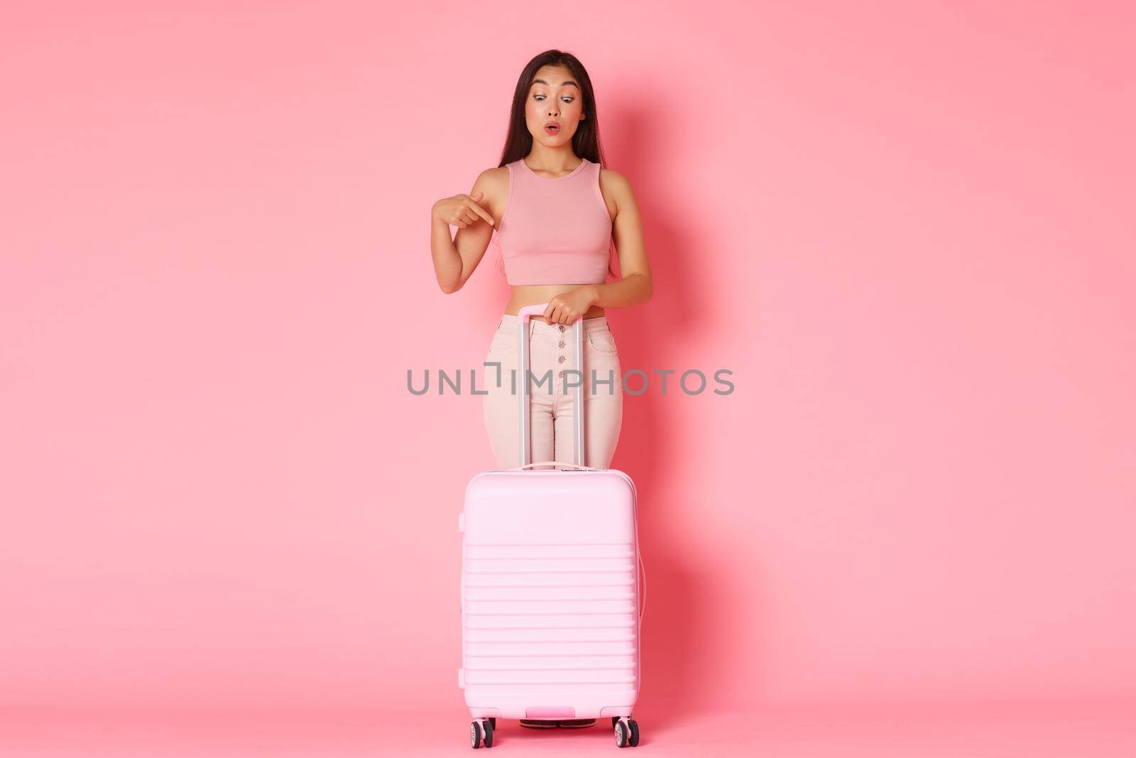 Travelling, holidays and vacation concept. Full-length of curious and interested asian tourist girl in summer clothes, holding suitcase, pointing finger down wondered, pink background by Benzoix