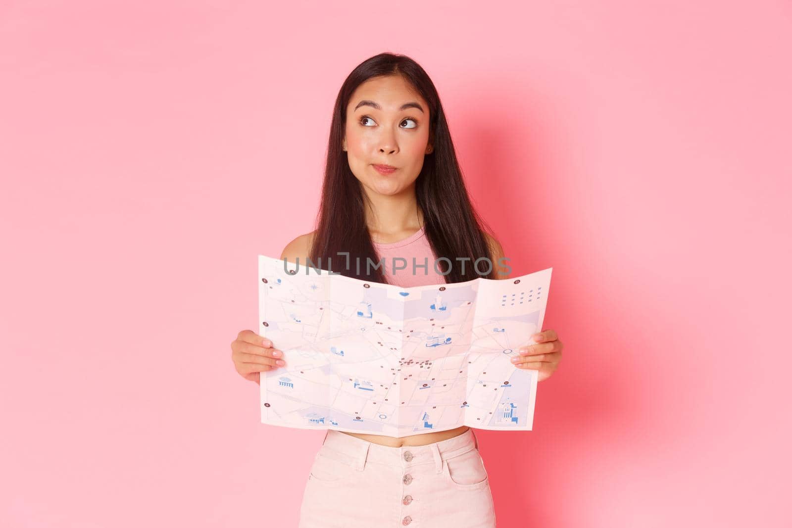 Travelling, lifestyle and tourism concept. Portrait of thoughtful attractive asian girl tourist looking upper left corner and thinking where to go while holding map of city, traveller make decision by Benzoix