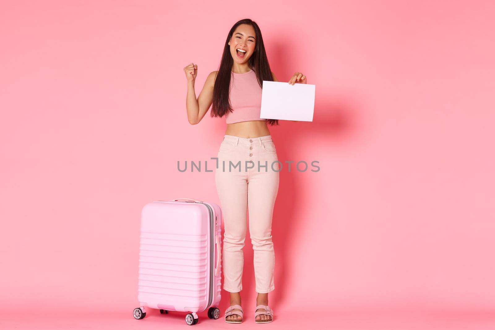 Travelling, holidays and vacation concept. Full-length of cheerful successful asian girl got opportunity travel abroad, standing with suitcase, fist pump in rejoice, holding piece of paper.