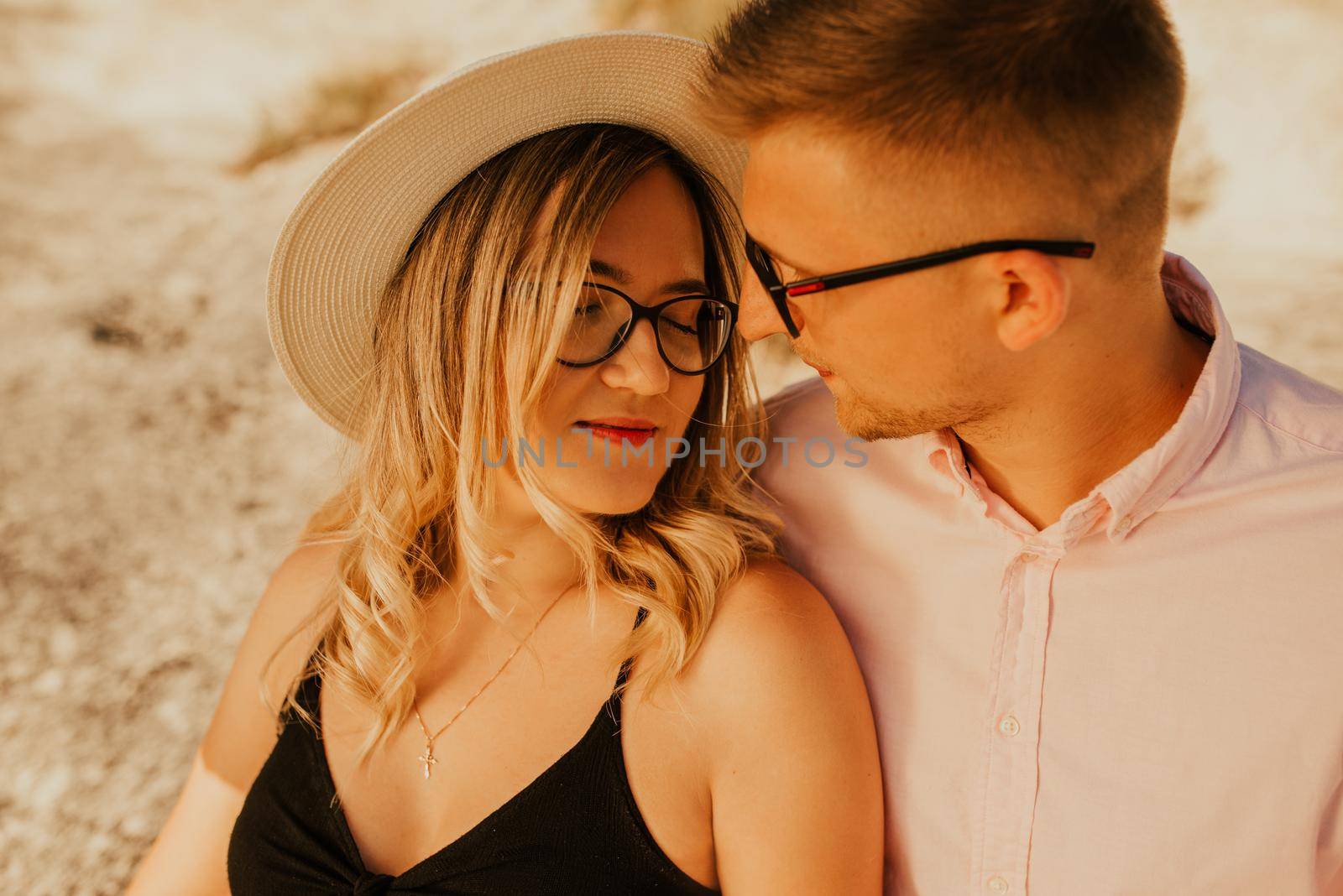 A man and a woman in a hat are sitting on a large stone rock. A couple of fair-haired fair-skinned people in love are resting in nature in a field at sunset.