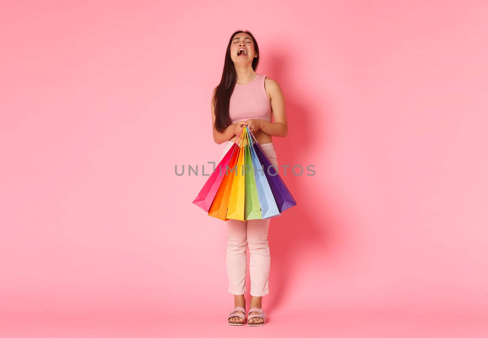 Full length portrait of crying and disappointed, whining asian girl complaining about not enough money, holding shopping bags and sobbing upset, standing pink background.