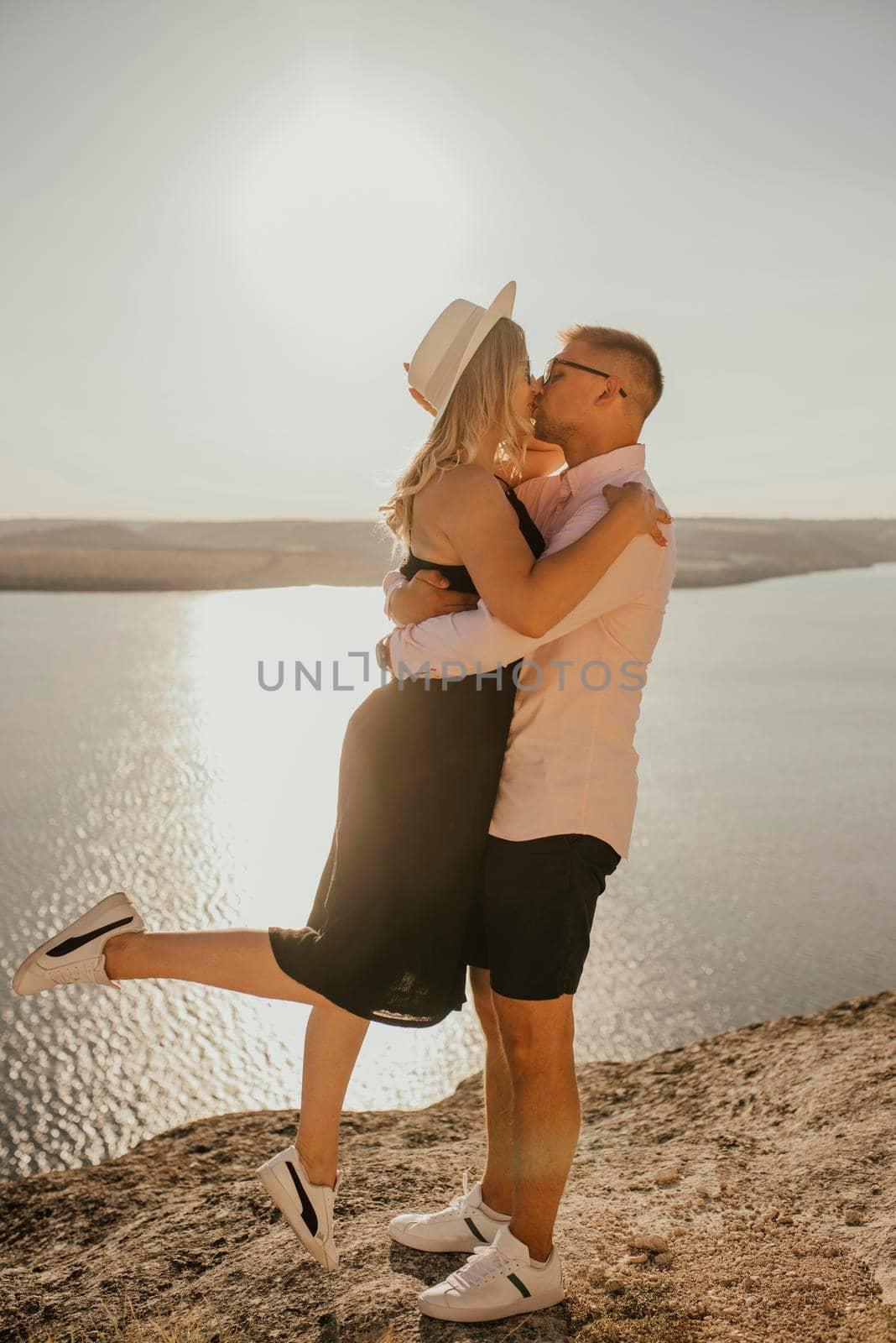 A man and a woman in a hat walk along the rocks above the cliff. A couple of fair-haired fair-skinned people in love are resting in nature in a field at sunset.