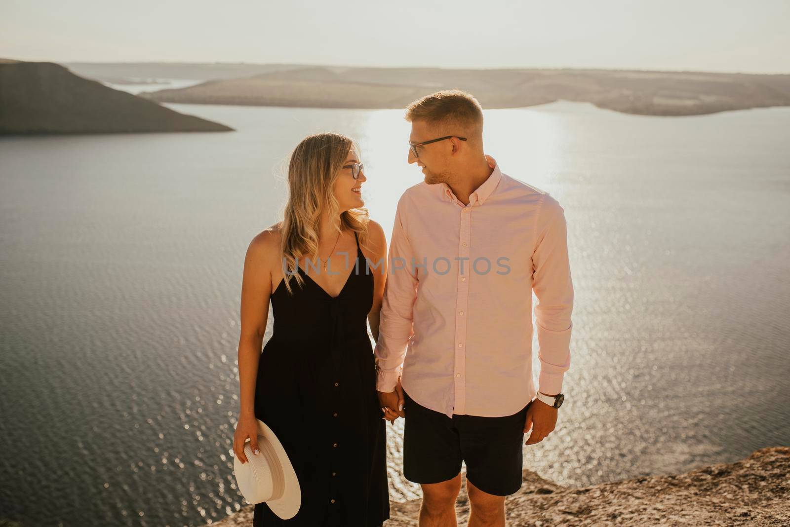 A man and a woman in love walk on a mountain above a large lake by the sea at sunset. A couple of fair-haired fair-skinned people in love are resting in nature in a field
