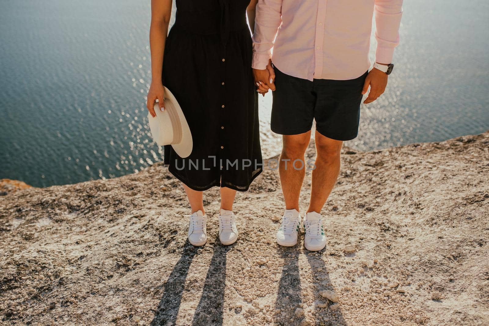 Male and female legs in white sneakers on a rock against the background of the sea. A couple of fair-haired fair-skinned people in love are resting in nature in a field at sunset.