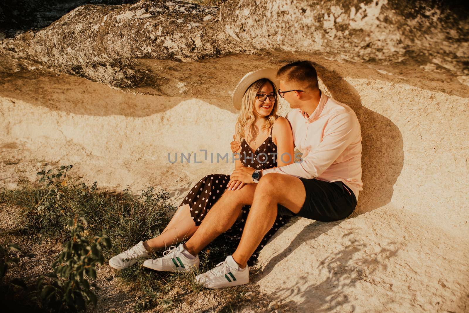 A man and a woman in a hat are sitting on a large stone rock. A couple of fair-haired fair-skinned people in love are resting in nature in a field at sunset.