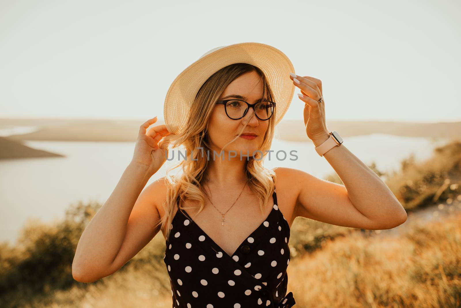 young woman in hat in dress with polka dots stands in middle of meadow on cliff above the sea at sunset