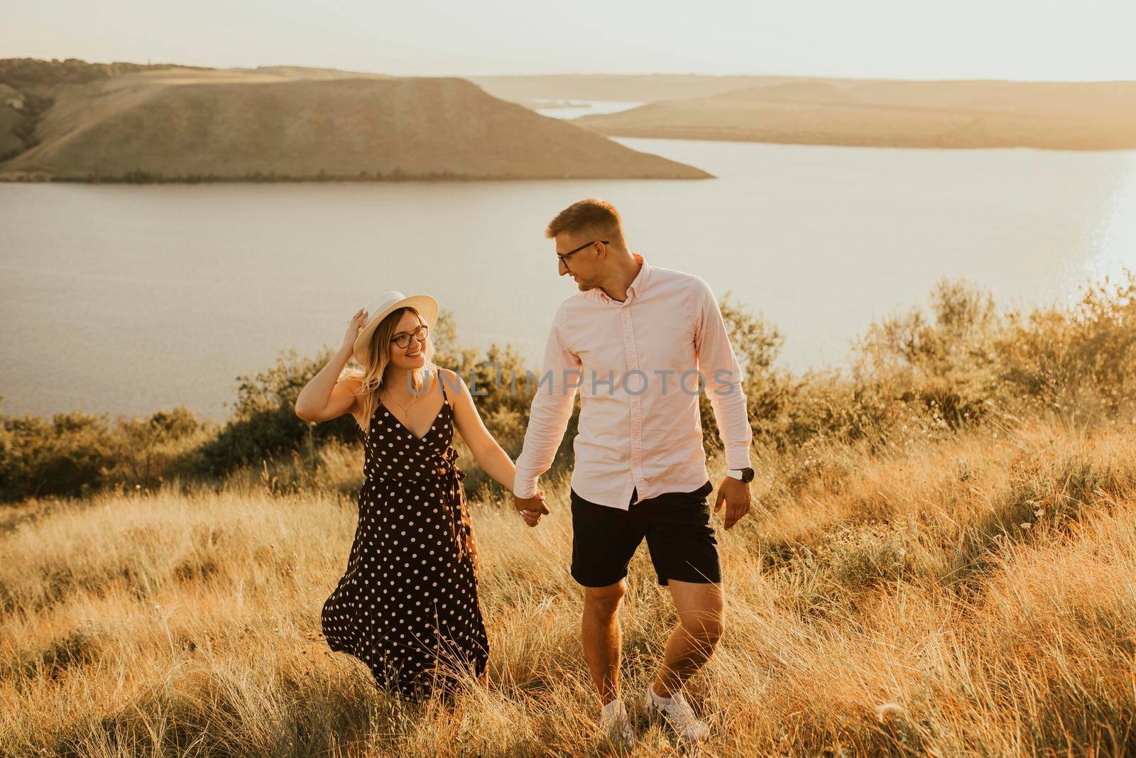 young man and woman walking in the meadow at sunset in summer near the lake. A couple of fair-haired fair-skinned people in love are resting in nature in a field at sunset.