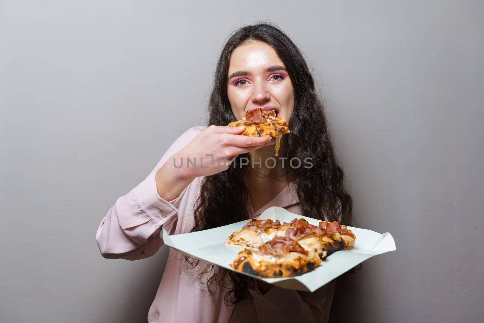 Girl courier eating pinsa pizza romana gourmet italian cuisine on grey background. Holding scrocchiarella traditional dish. Food delivery from pizzeria. Pinsa with meat, arugula, olives, cheese.