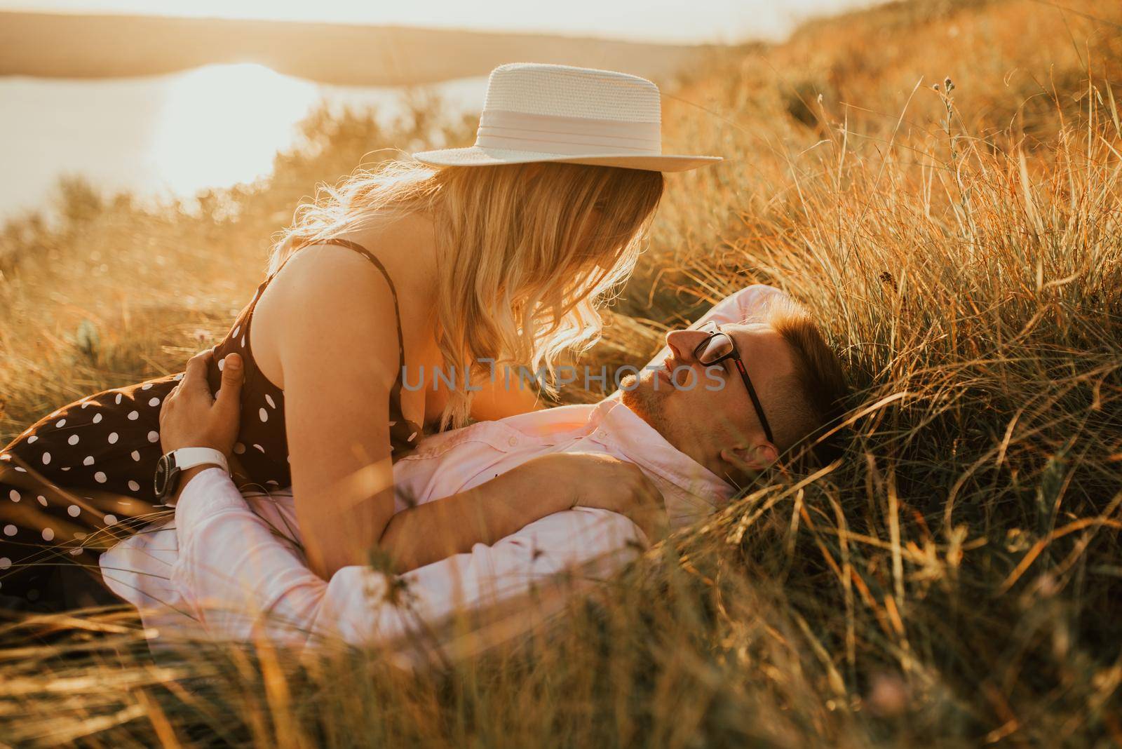 A man and a woman in a hat lie on the ground in the middle of the grass. A couple of fair-haired fair-skinned people in love are resting in nature in a field at sunset.