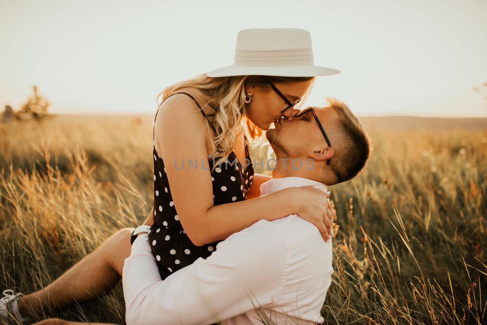 A girl in hat sits in hands guy on ground in middle of tall grass in meadow. couple of fair-haired fair-skinned people in love are resting in nature in field at sunset.