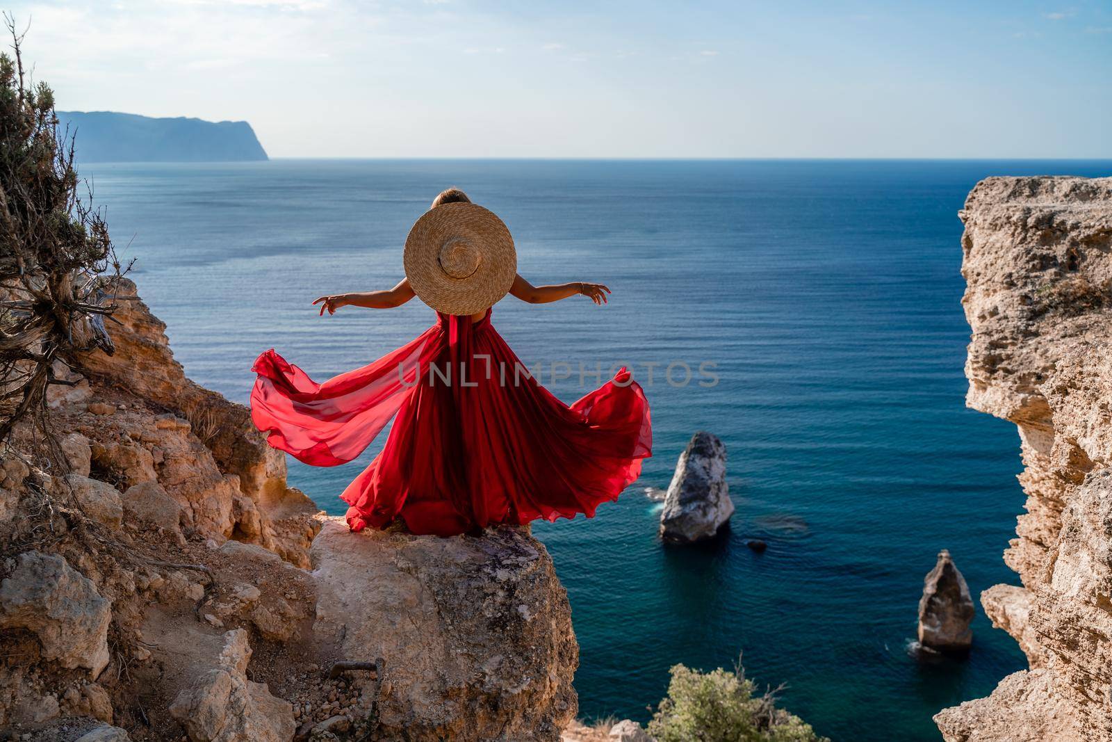 A woman in a flying red dress fluttering in the wind and a straw hat against the backdrop of the sea