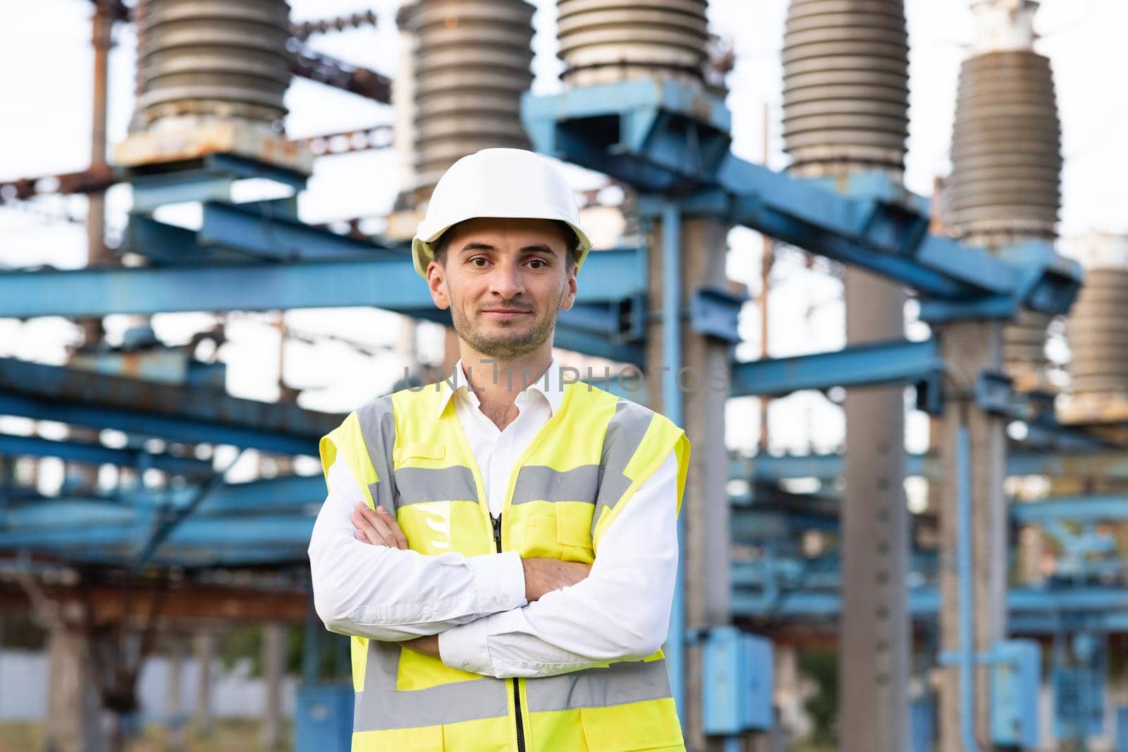 Portrait of young smiling asian electric worker with arms folded at power site. Portrait of engineer worker in hard hat standing at high voltage power station.