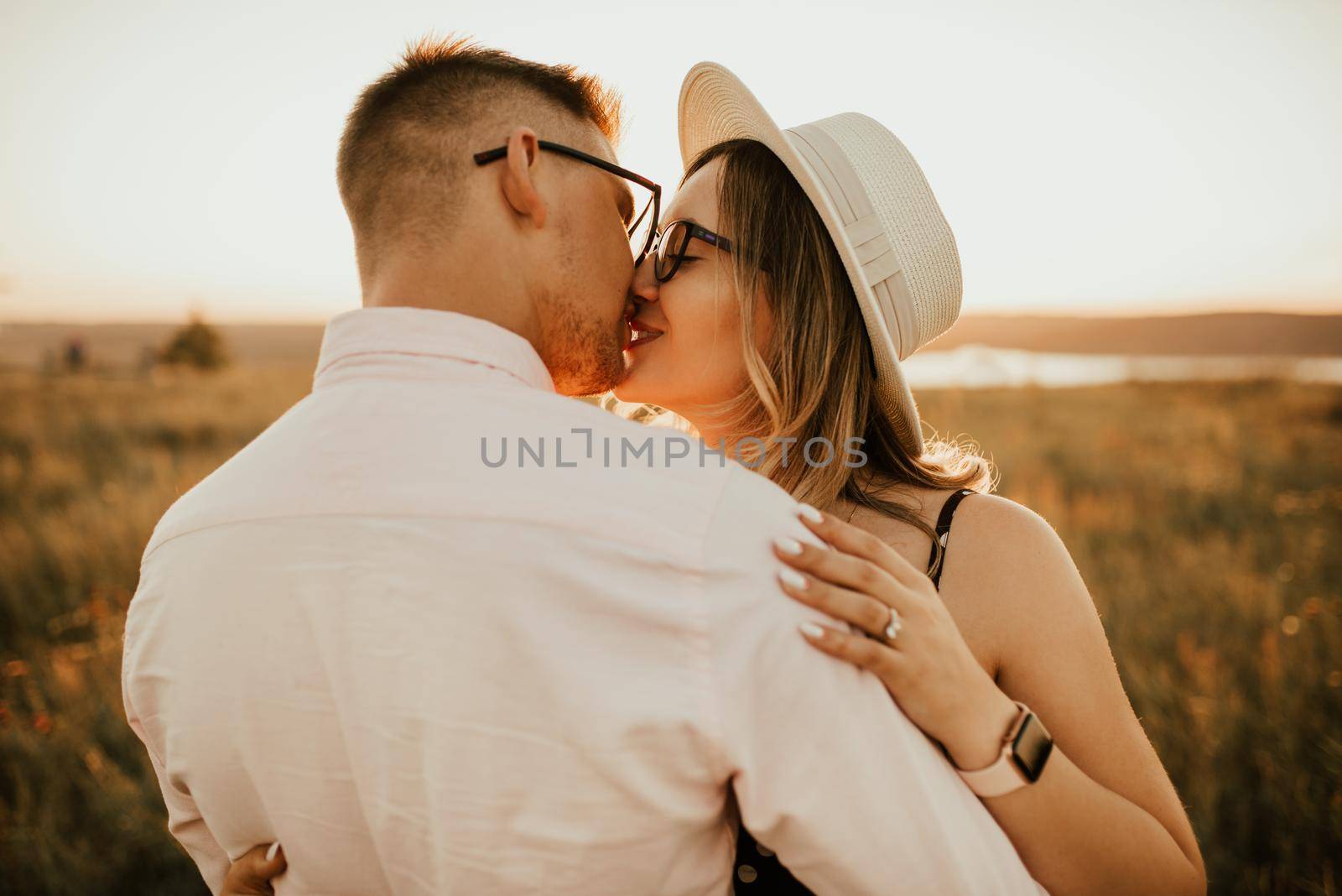 A man with a woman in a hat hug and kiss in the tall grass in the meadow. A couple of fair-haired fair-skinned people in love are resting in nature in a field at sunset.