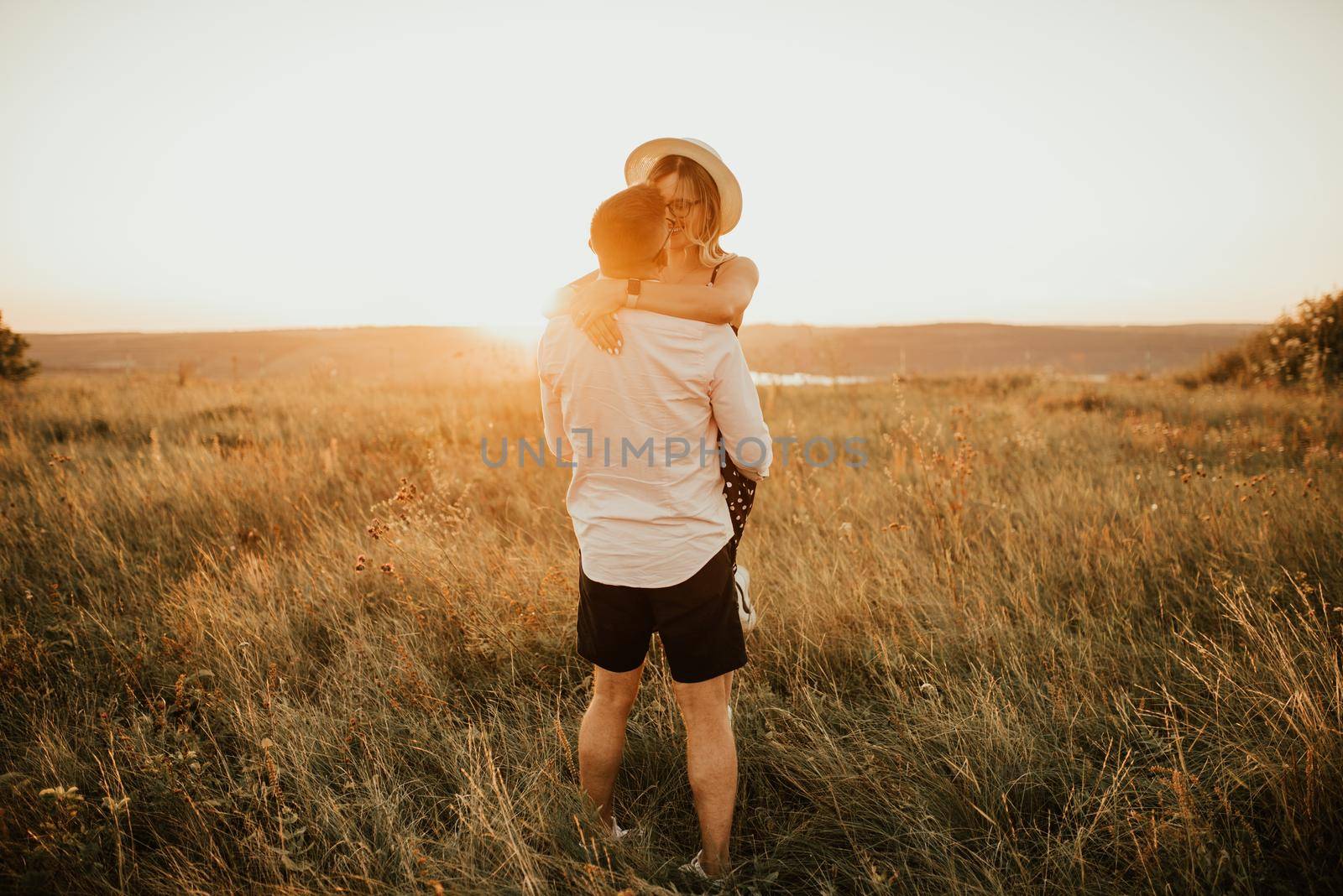 A man with a woman in a hat hug and kiss in the tall grass in the meadow. A couple of fair-haired fair-skinned people in love are resting in nature in a field at sunset.