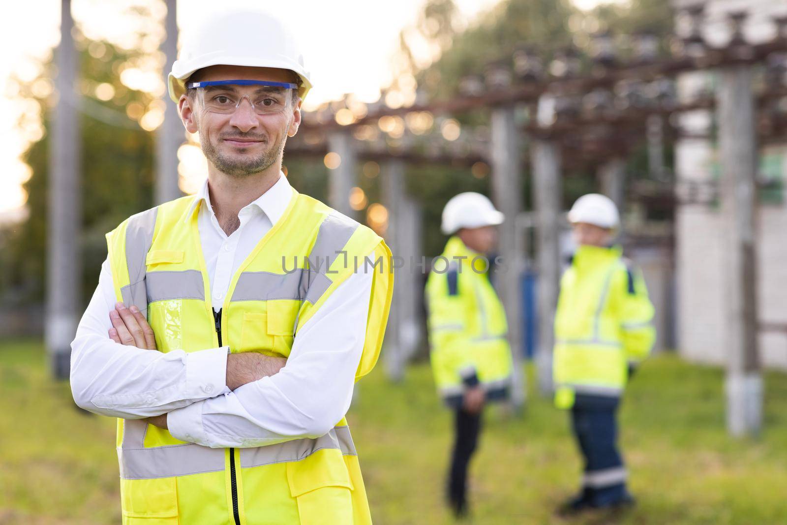 Portrait of male engineer technologist standing among high voltage electrical lines with her arms crossed over her chest. Man in protective helmet and uniform smiling and looking at camera by uflypro